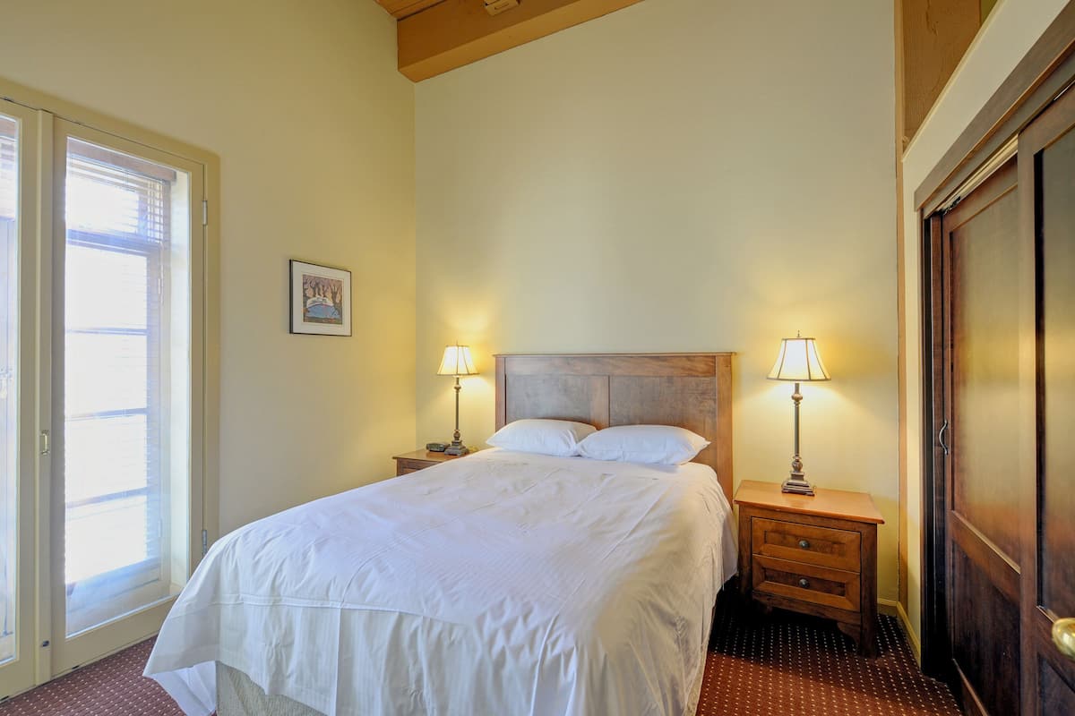 A neatly made bed with white linens, two bedside tables with lamps, a wooden headboard, a door, and a window with blinds.