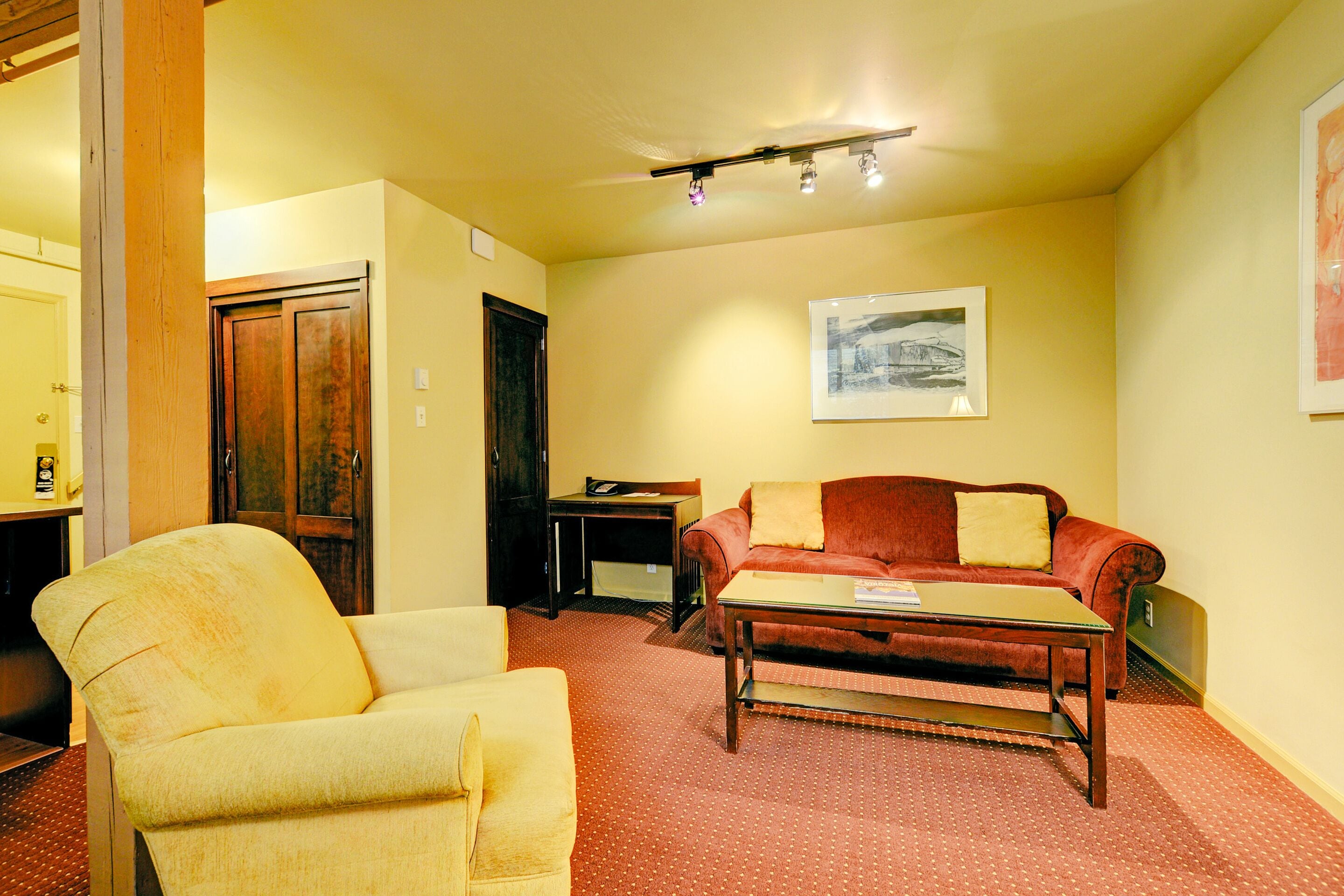 A living room with a yellow armchair, a red sofa, and a wooden coffee table. There is a door and a framed picture on the wall.