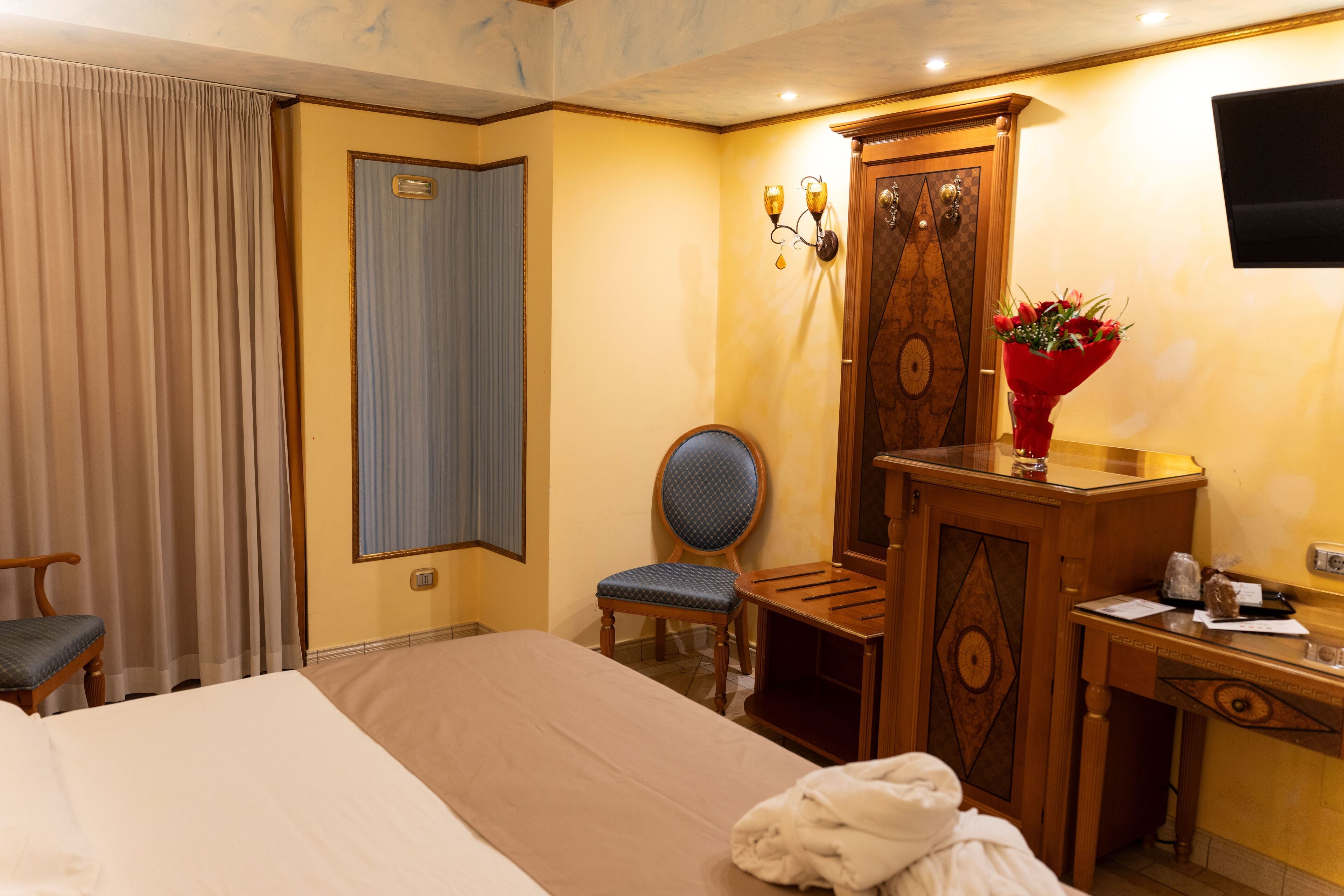 A hotel room with a bed, a desk, a chair, a wooden wardrobe, a television, and a vase with flowers.