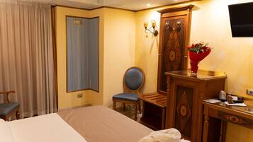 A hotel room with a bed, a desk, a chair, a wooden wardrobe, a television, and a vase with flowers.