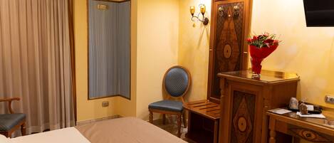 A hotel room with a bed, a desk, a chair, a wooden wardrobe, a television, and a vase with flowers.