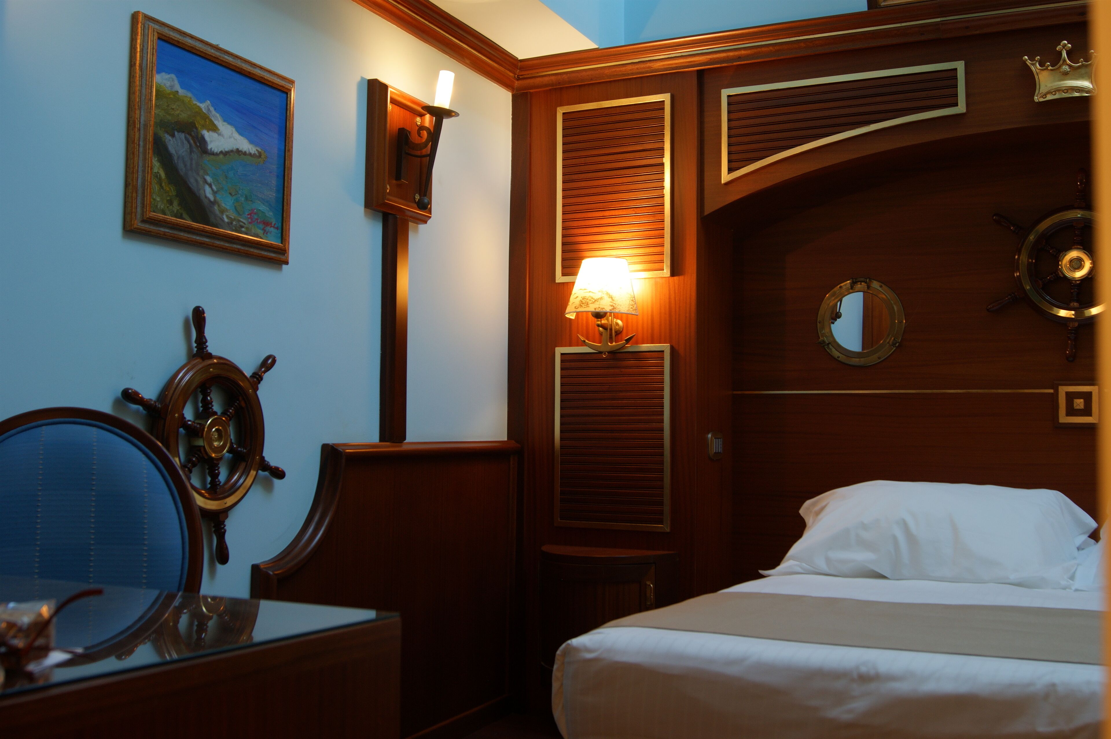 Une chambre d’hôtel comprenant un lit, un bureau avec une chaise, un gouvernail de bateau, un tableau représentant un paysage côtier et une lampe fixée au mur.