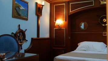 Une chambre d’hôtel comprenant un lit, un bureau avec une chaise, un gouvernail de bateau, un tableau représentant un paysage côtier et une lampe fixée au mur.