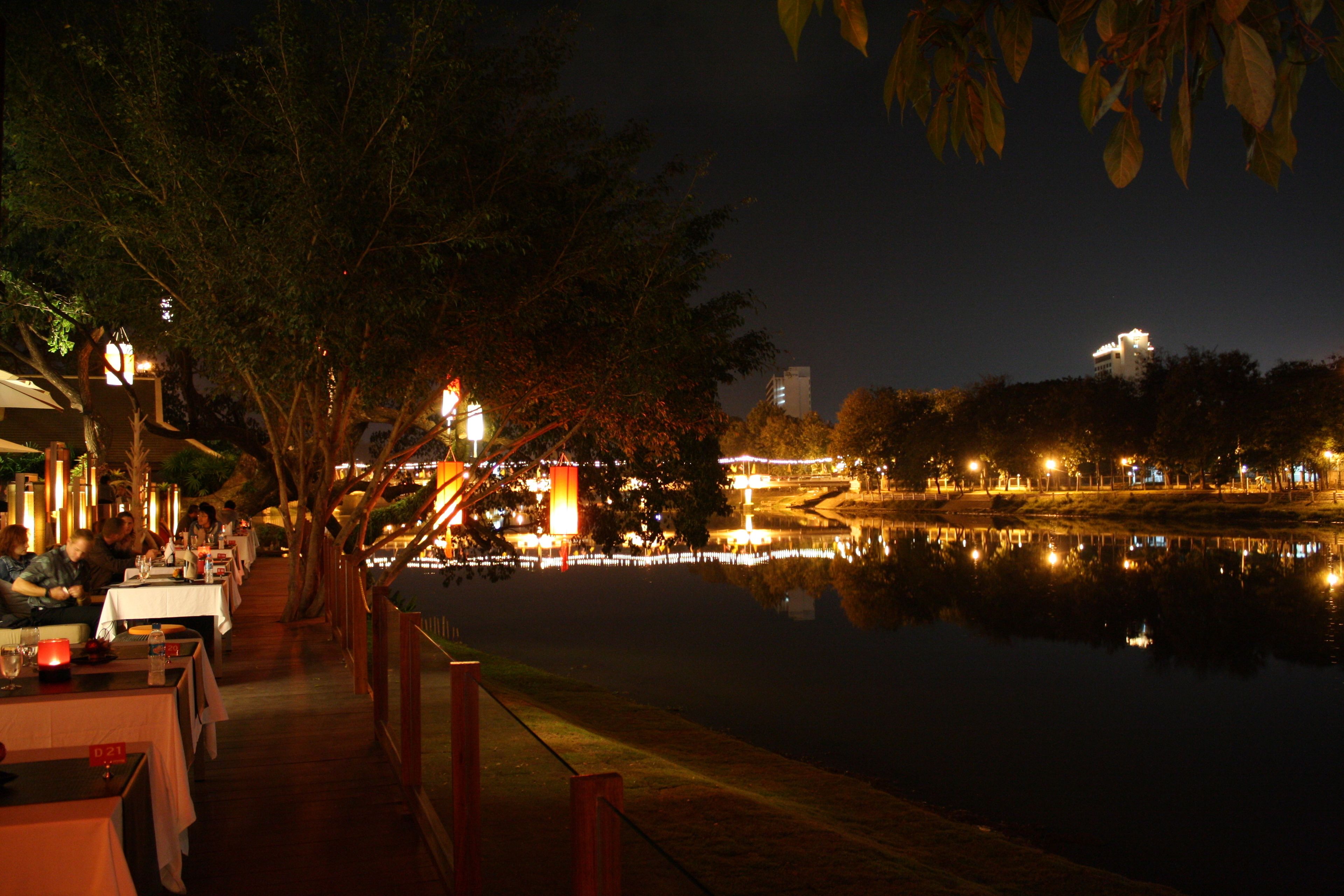 A riverside dining area with tables and chairs, a wooden deck, and a view of a cityscape across the water.