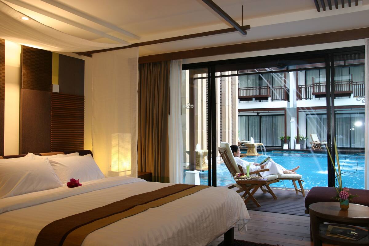 A hotel room with a large bed, a view of a swimming pool, and a balcony with lounge chairs.