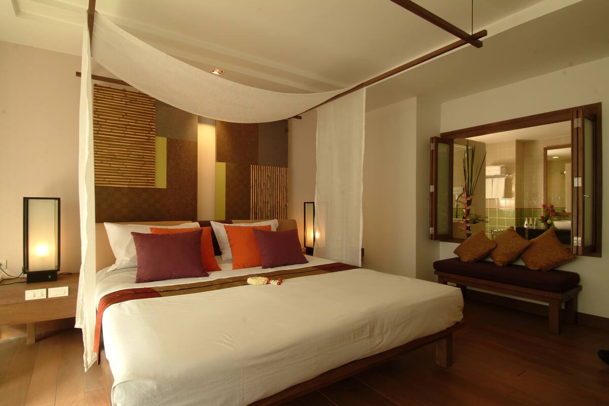 A spacious bedroom with a large bed, a bench, and a mirror reflecting the bathroom.