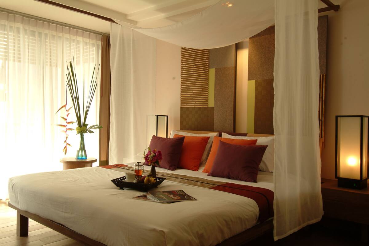 A hotel room with a large bed, a bedside table, a nightstand, a lamp, a vase with flowers, and a window with curtains.