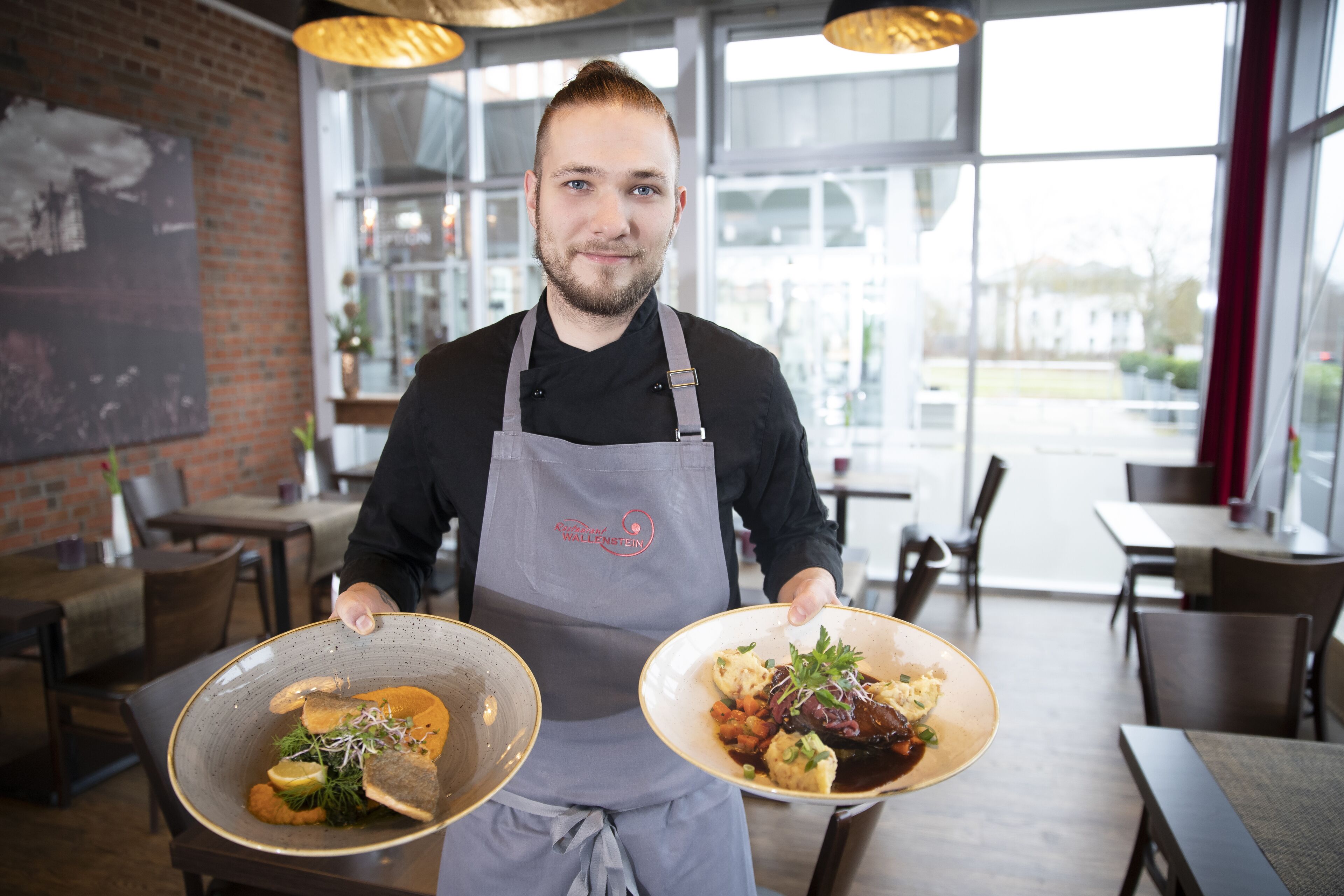 Een persoon die in een restaurant met bakstenen muren en grote ramen twee borden vasthoudt.
