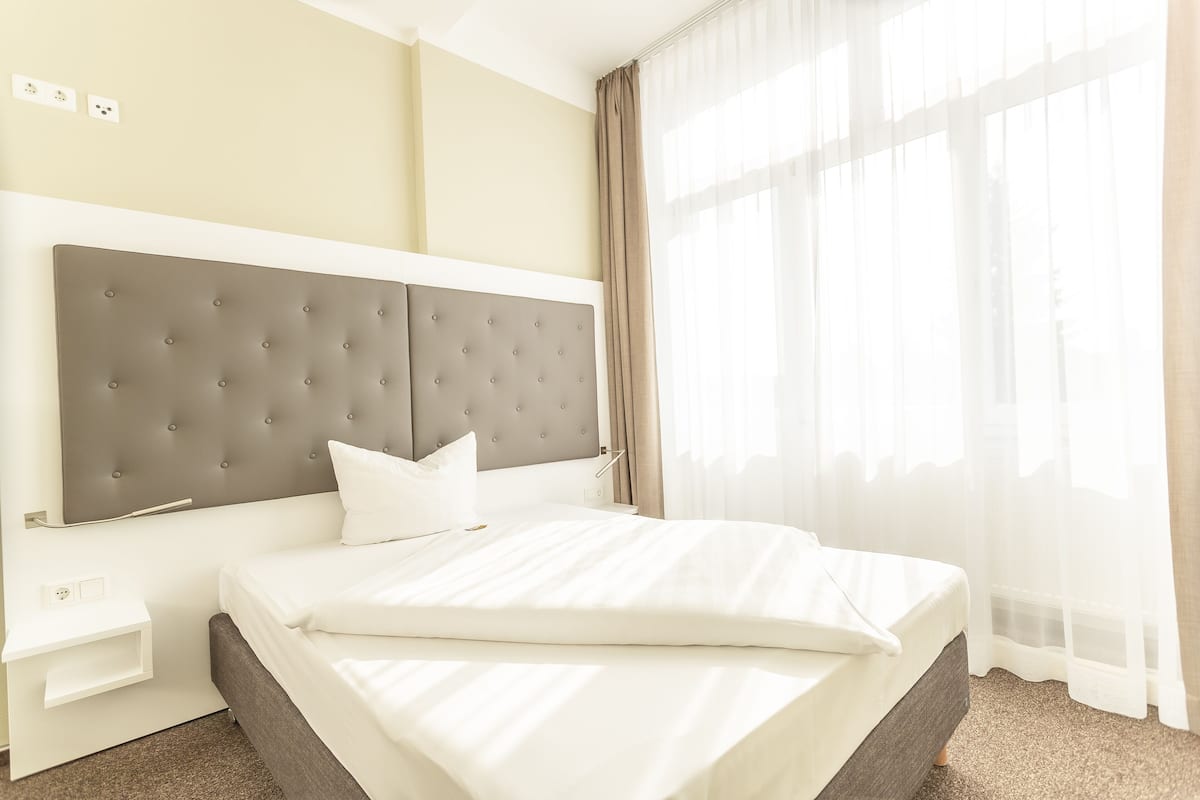 Una cama bien hecha con un cabecero capitonado, una mesita de noche y una ventana con cortinas translúcidas.