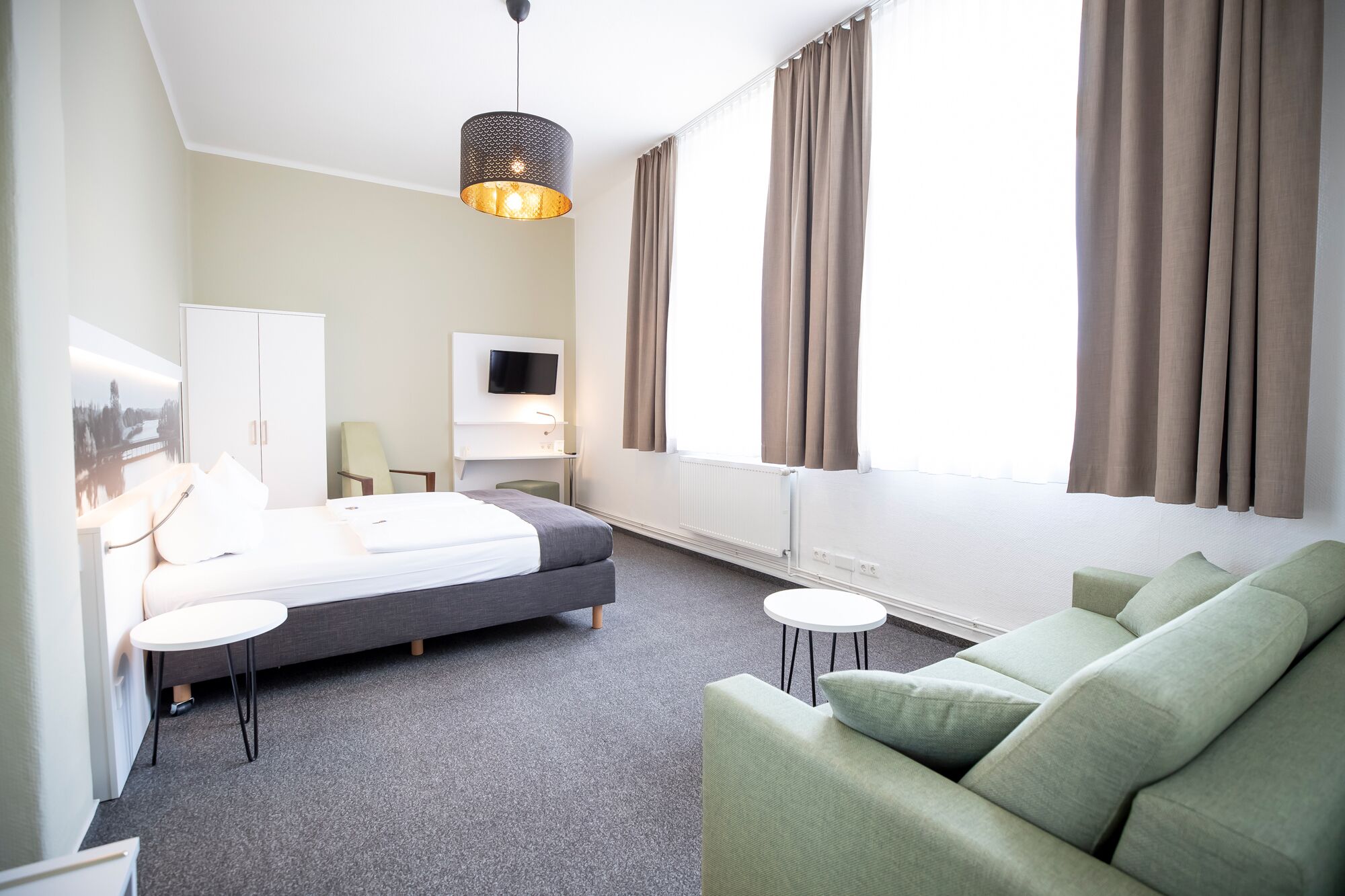 Habitación de hotel con una cama, un sofá verde, una mesita pequeña, un televisor y una ventana con cortinas.