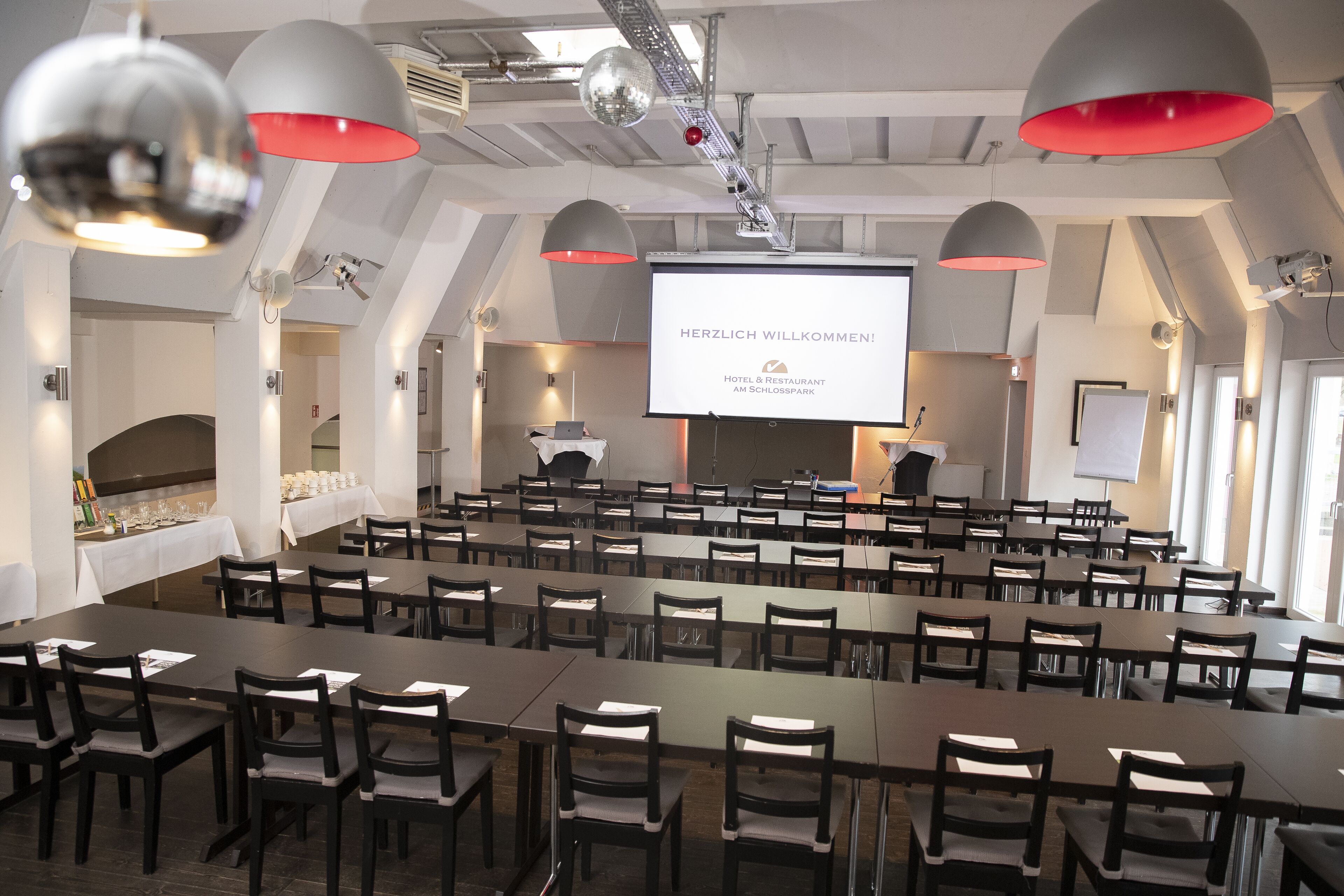 A conference room with tables and chairs, a projector screen displaying a welcome message, and a stage with a podium.