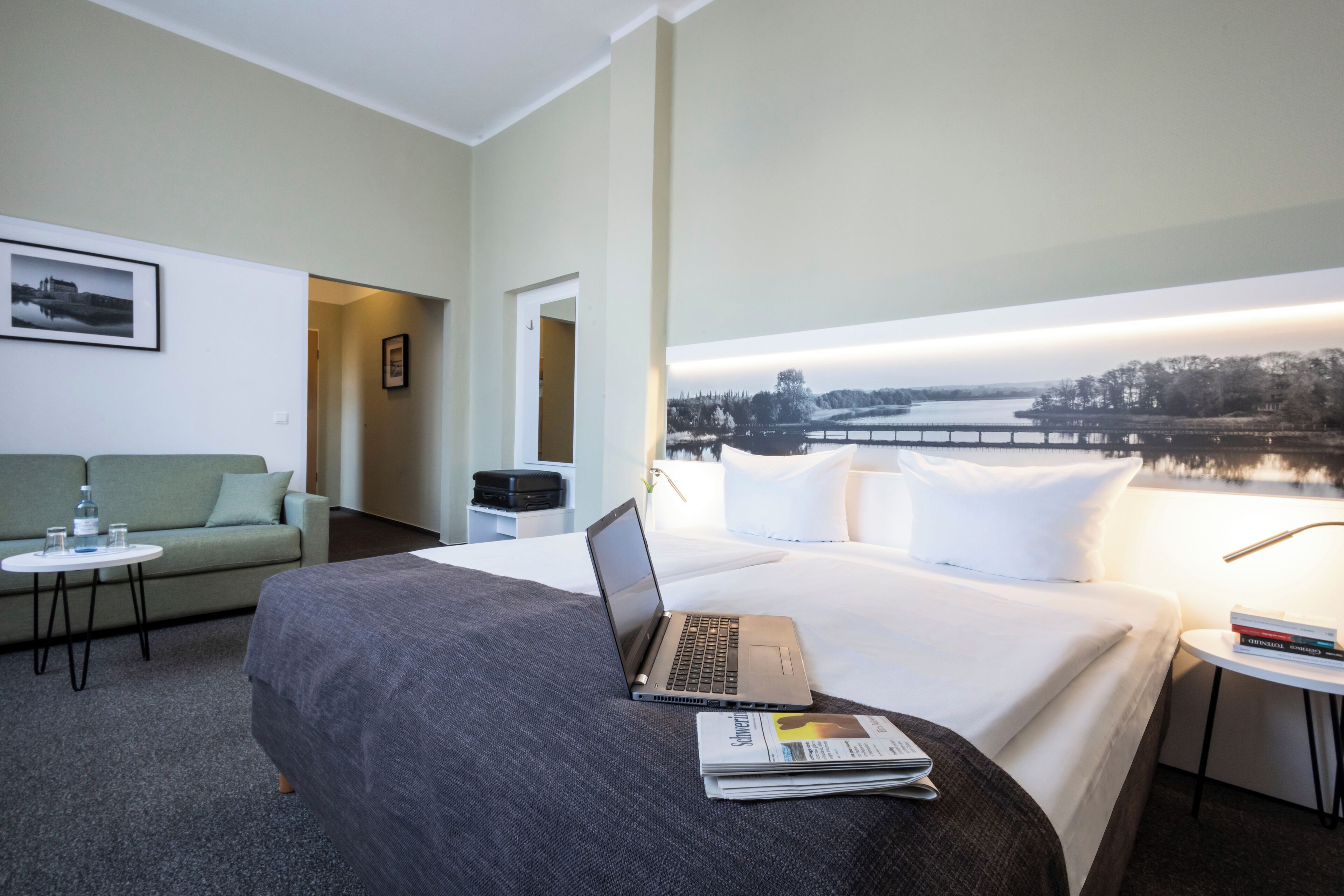 Habitación de hotel con cama, una laptop, una mesita con un libro, un sofá y vistas a un lago.