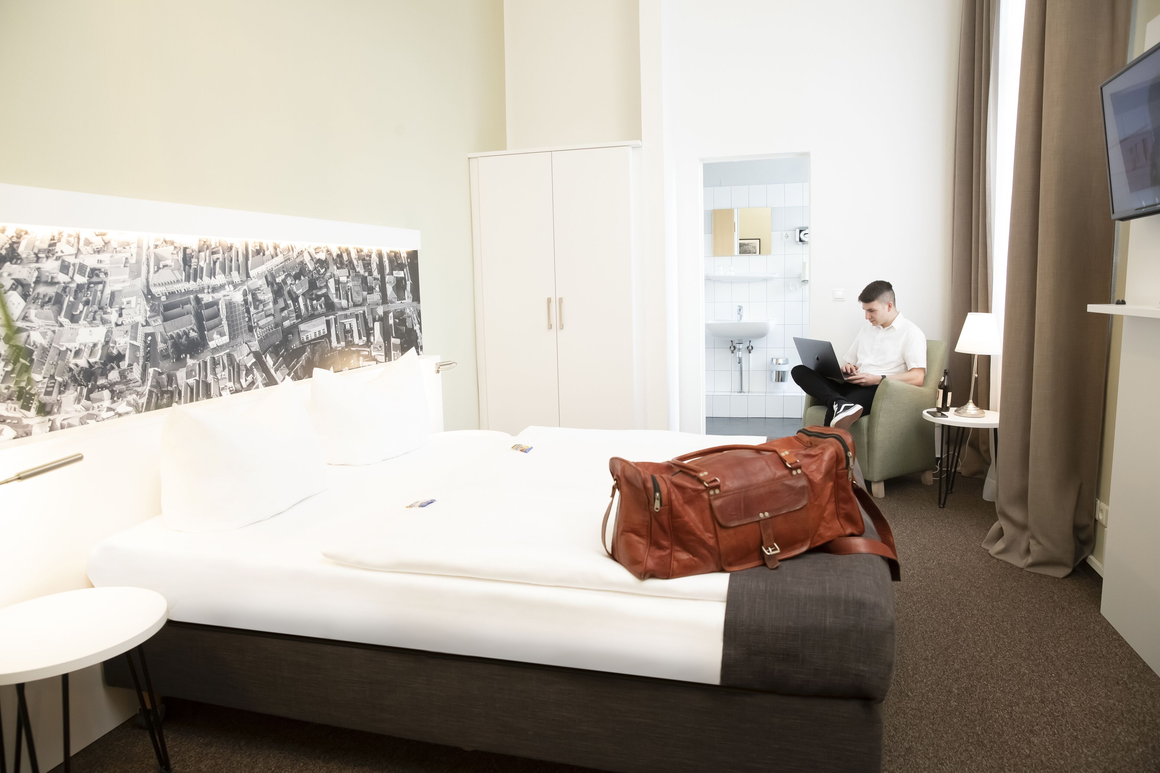 A hotel room with a bed, a desk, a chair, a wardrobe, a TV, and a bathroom visible through an open door.