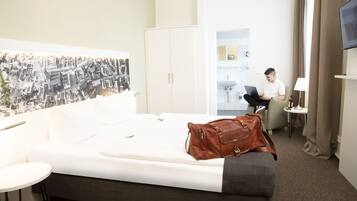 A hotel room with a bed, a desk, a chair, a wardrobe, a TV, and a bathroom visible through an open door.