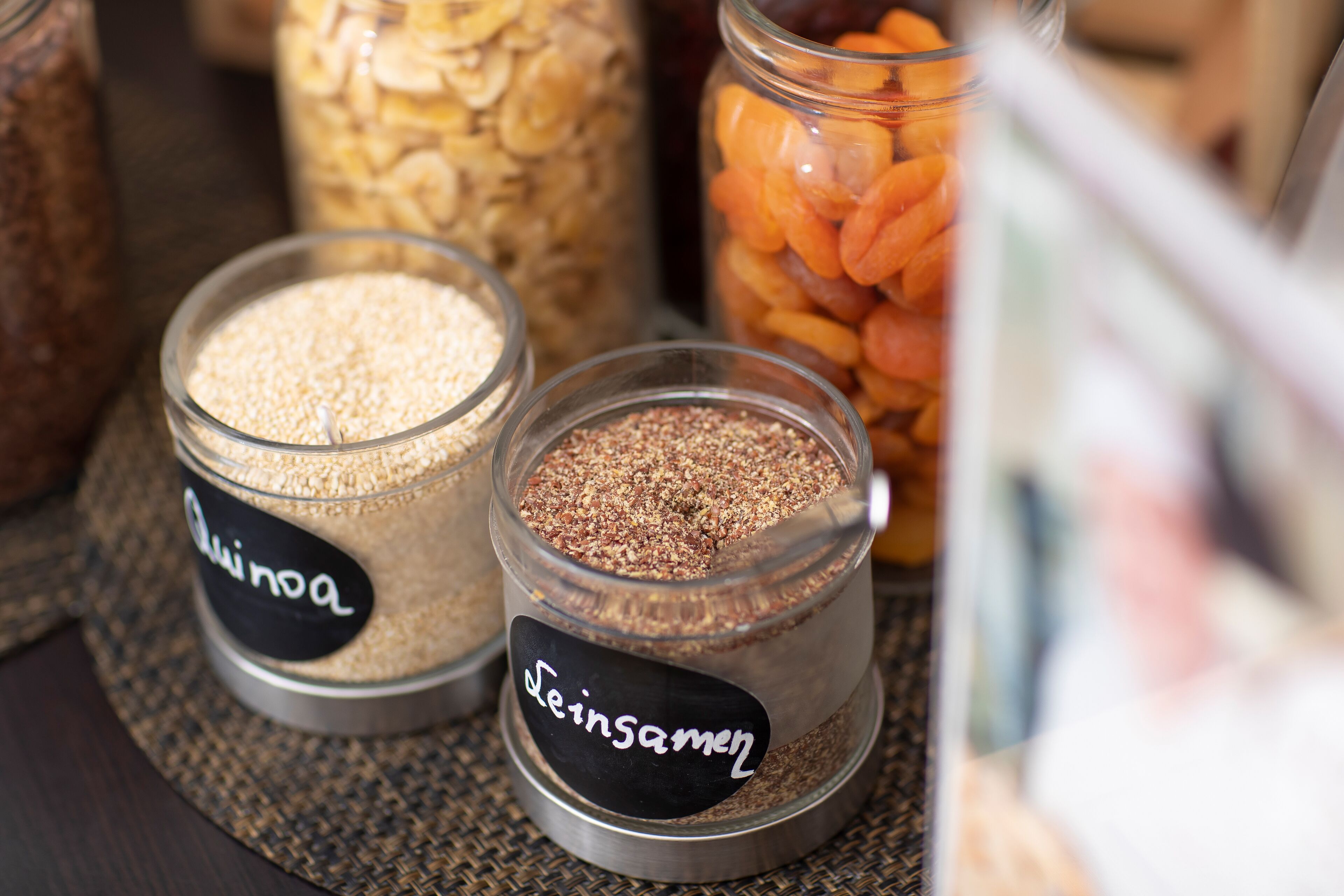 Dos frascos de vidrio etiquetados con “Quinoa” y “Seinsaemen” sobre una estera tejida.