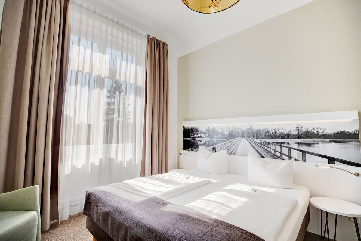 Habitación de hotel con una cama grande, una mesita redonda pequeña, una silla verde y un cuadro de un puente sobre el agua en la pared.