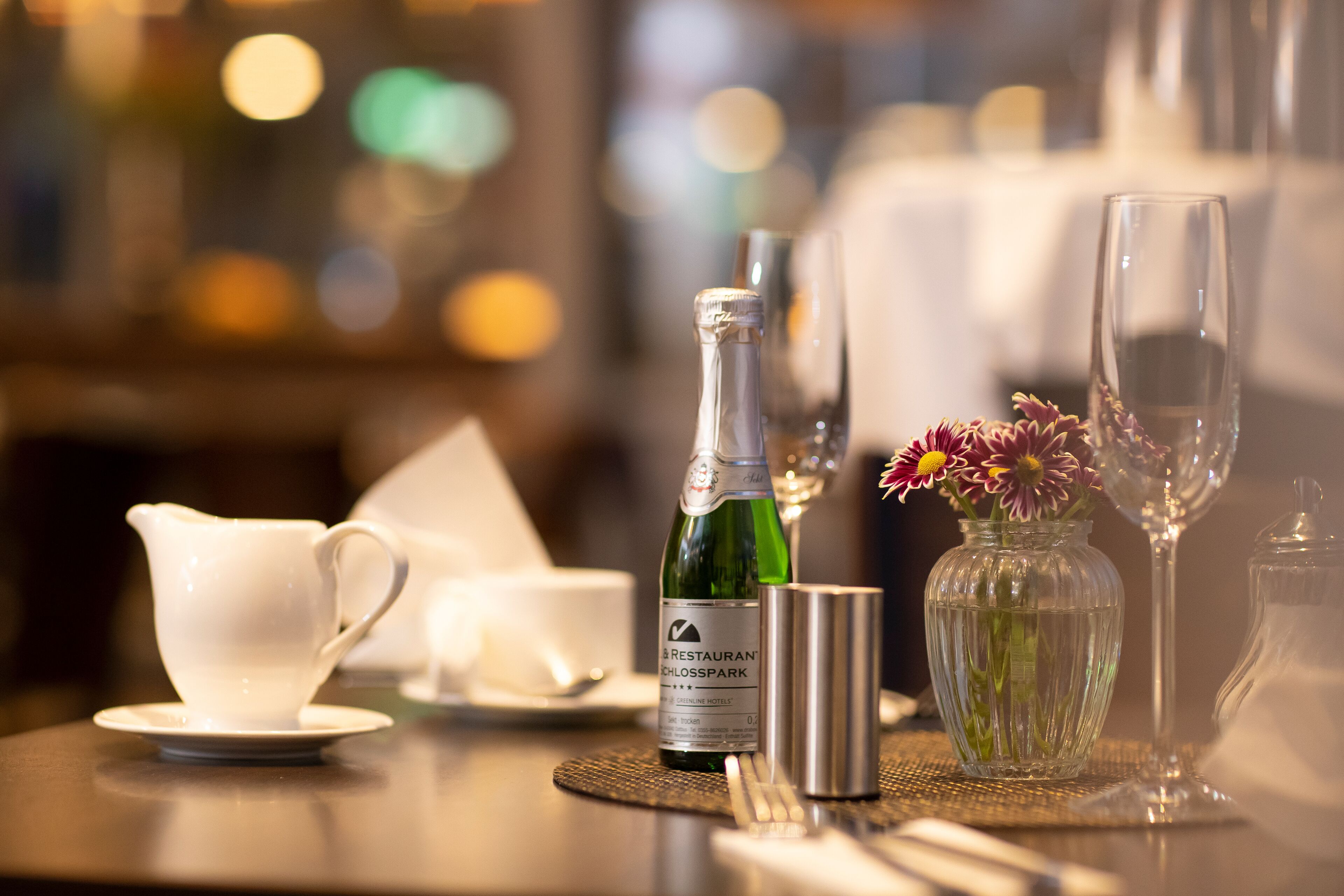 A dining table with a bottle of wine, a vase with flowers, a white cup, a silver cup, and a silver pitcher.