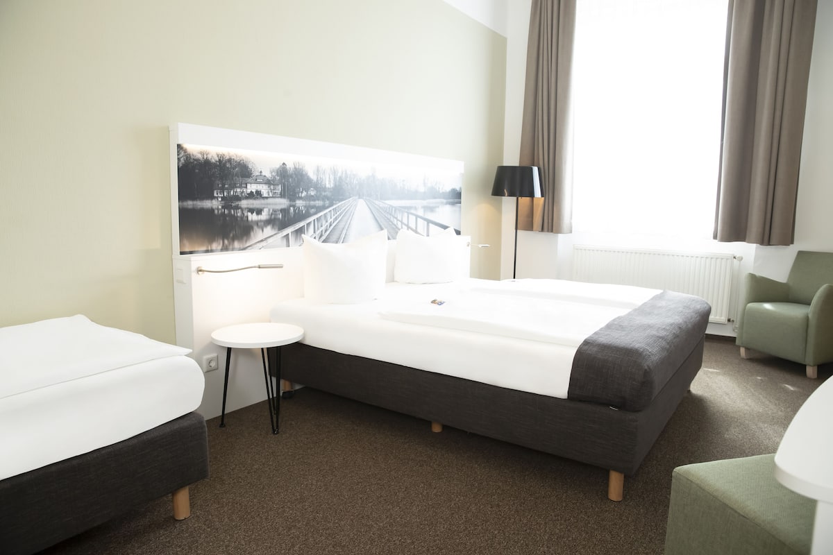 Habitación de hotel con una cama grande, una mesita redonda pequeña, una silla y un cuadro de un puente sobre el agua en la pared.