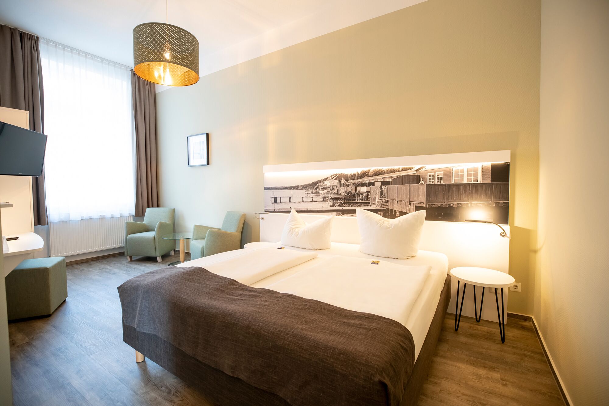 Een hotelkamer met een bed, twee fauteuils, een klein tafeltje, een bureau en een groot raam met gordijnen.
