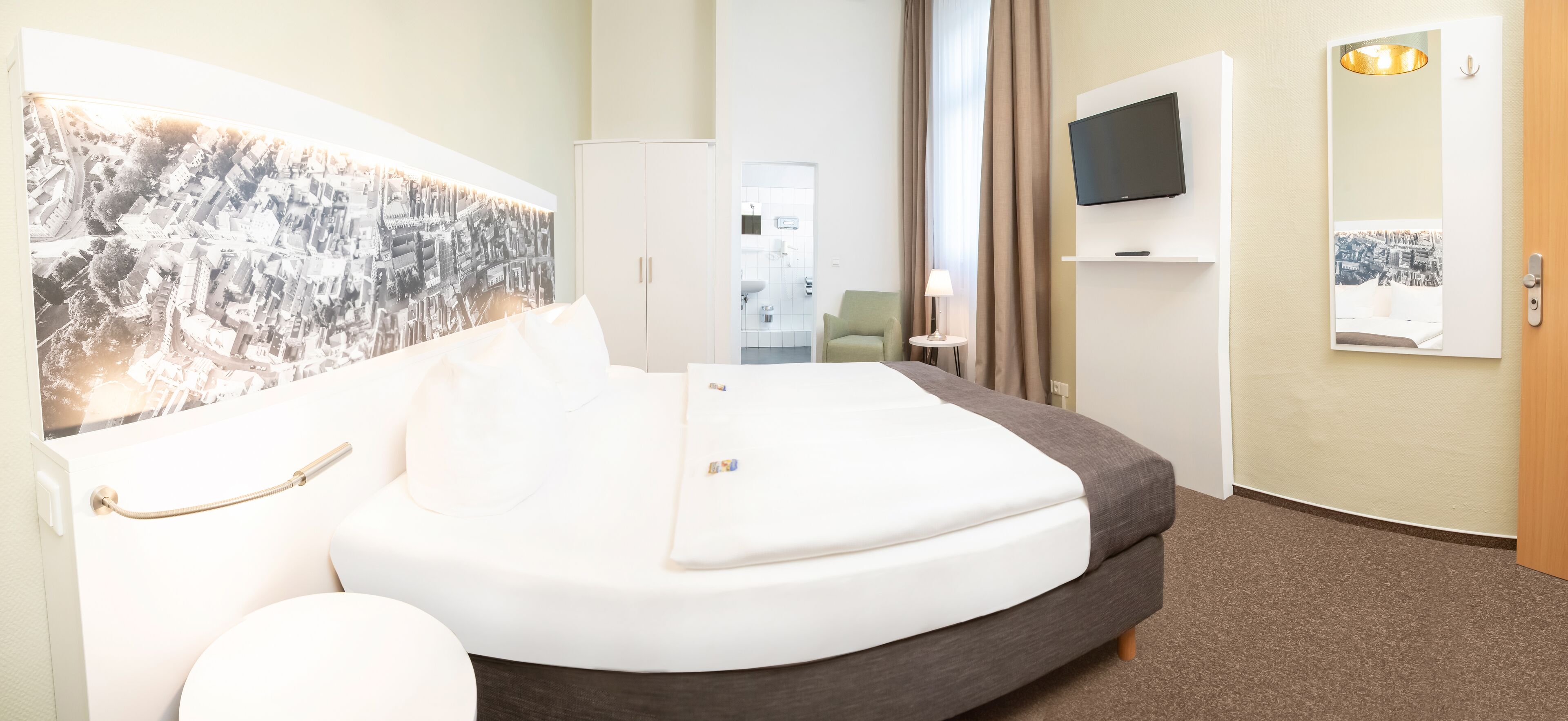 A modern hotel room with a large bed, a television, a mirror, and a cityscape artwork on the wall.