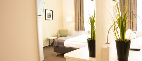 A hotel room with a bed, bedside table, potted plants, a lamp, and a window with curtains.