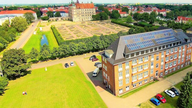 Un edificio grande con techo de paneles solares, rodeado por un césped verde y un estacionamiento con varios coches.