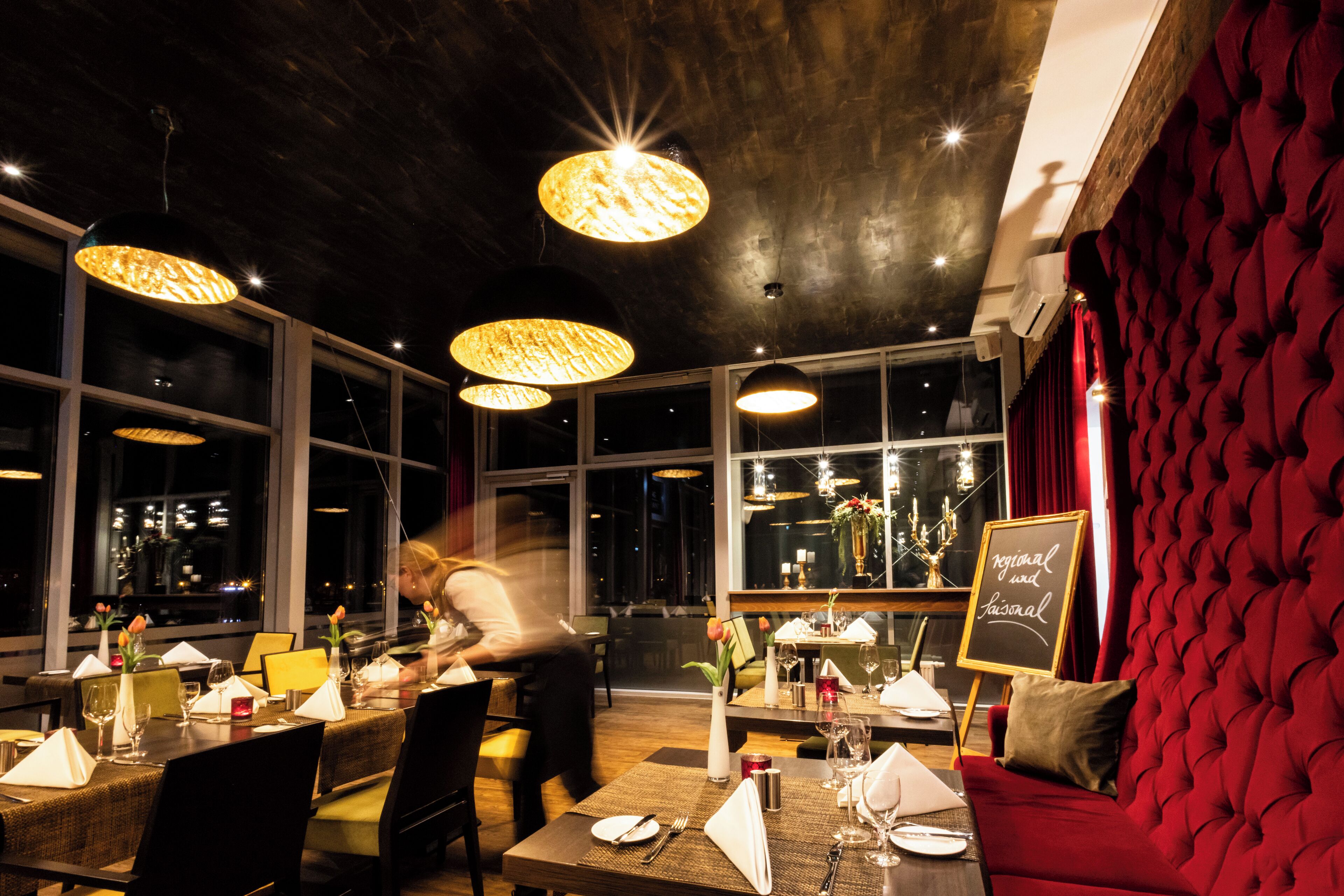 A restaurant with tables set for dining, large windows, and hanging light fixtures.
