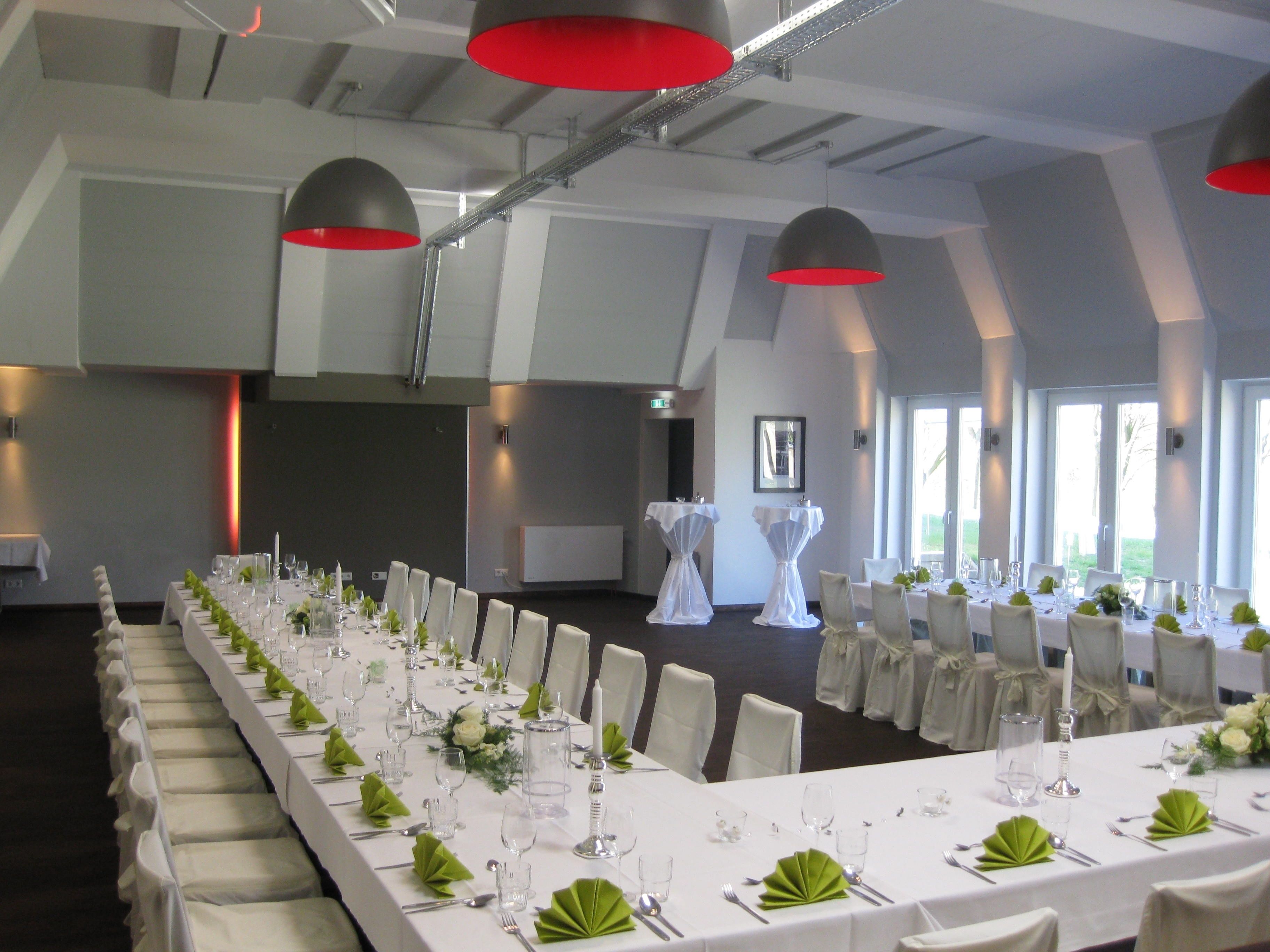 Une longue table de banquet, dressée pour un événement formel, avec des nappes blanches, des serviettes vertes et des luminaires suspendus rouges.