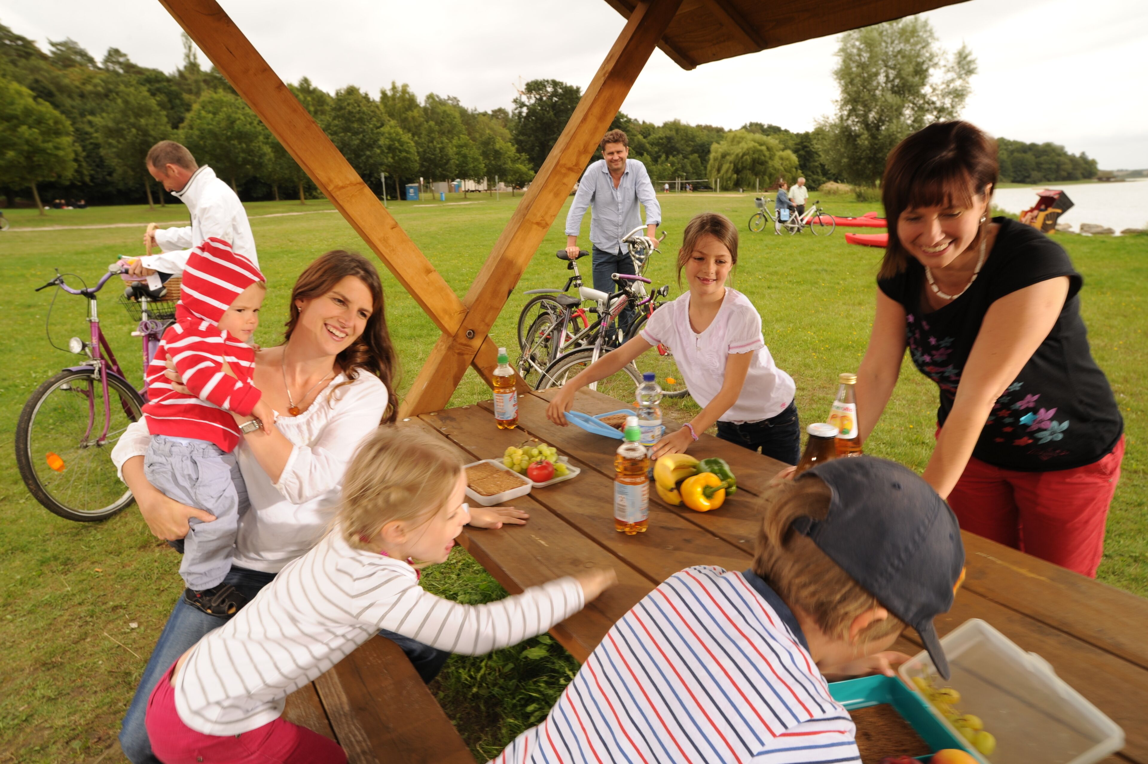 A group of people at a picnic table with food and drinks, outdoors near a body of water.