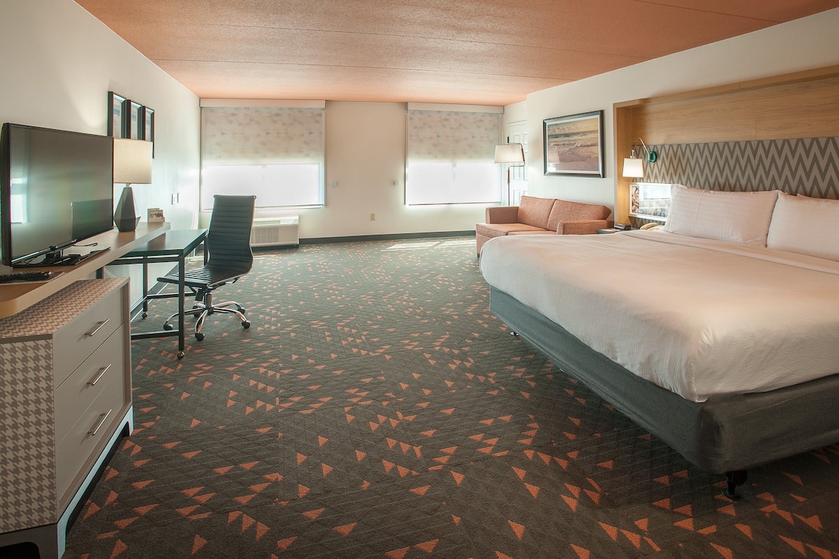 Una habitación de hotel con una cama, un escritorio con televisor, una silla y una alfombra con diseño geométrico.