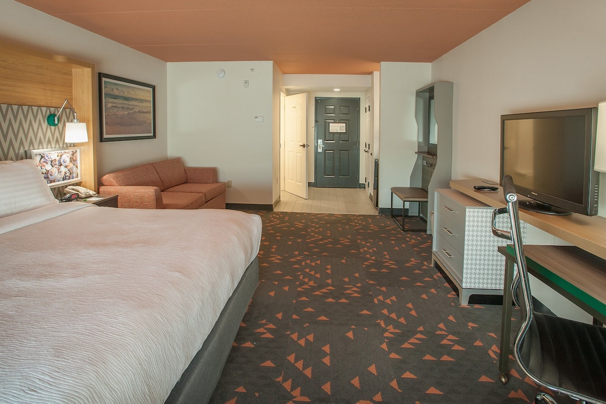 Habitación de hotel con cama, sofá, escritorio con televisor, silla y lámpara de escritorio.