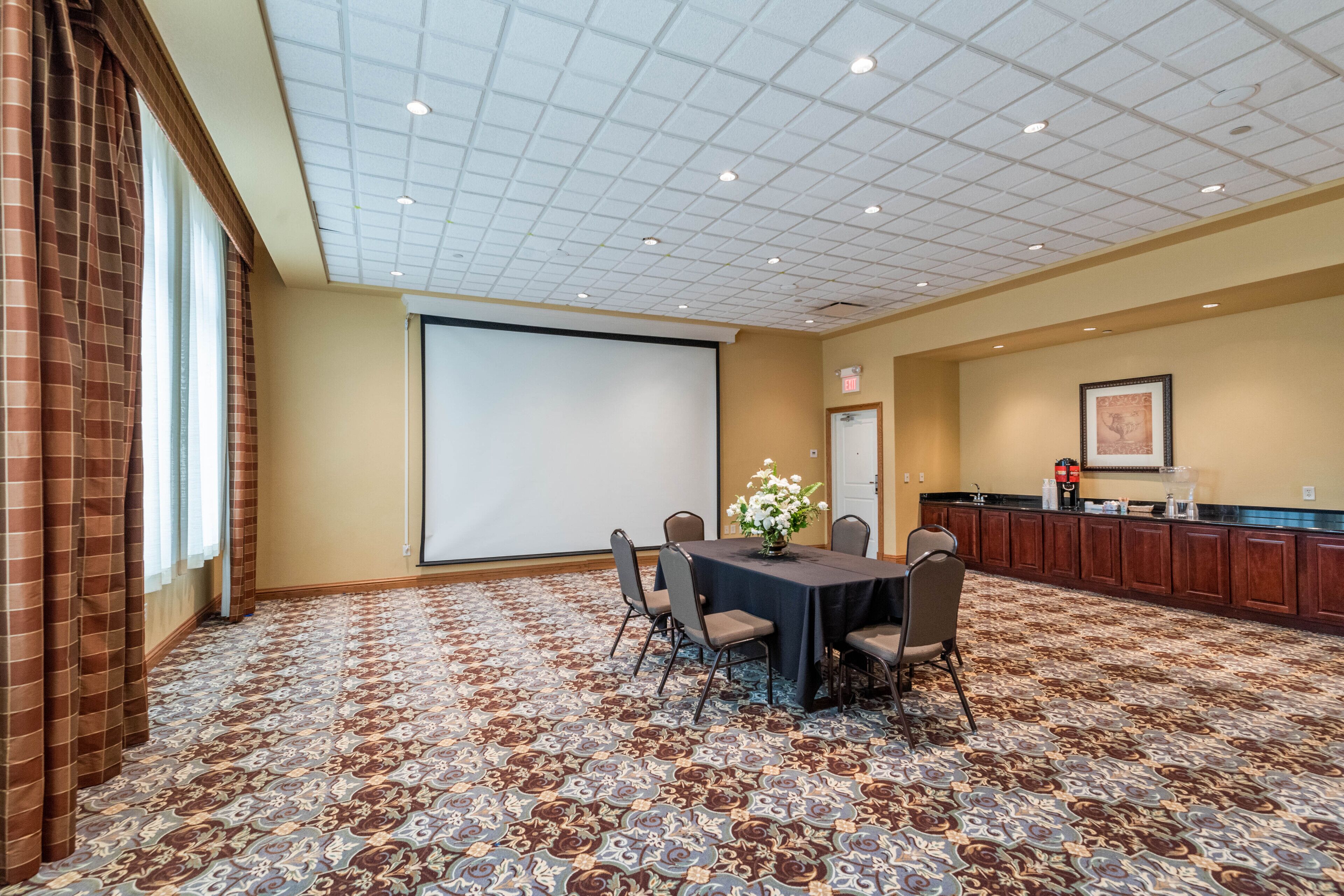 A conference room with a carpeted floor, a projector screen, a table with chairs, a flower vase, and a kitchenette area.