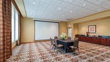 A conference room with a carpeted floor, a projector screen, a table with chairs, a flower vase, and a kitchenette area.