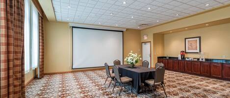 A conference room with a carpeted floor, a projector screen, a table with chairs, a flower vase, and a kitchenette area.