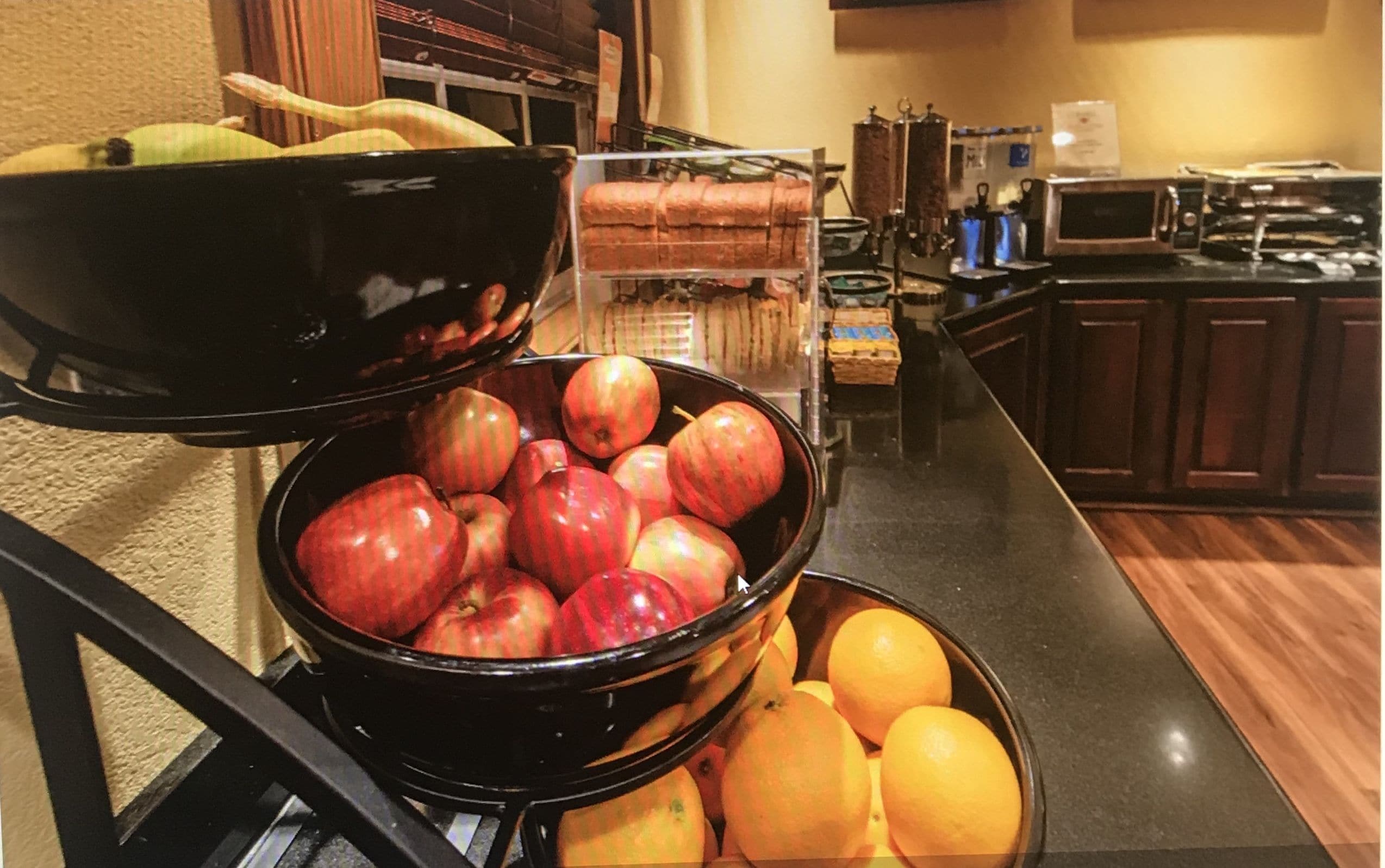 A kitchen area with a bowl of apples and oranges, a microwave, and a bread rack.