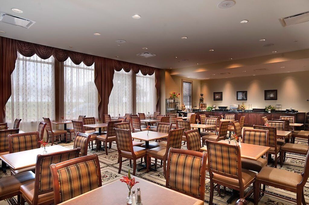 A hotel dining area with multiple tables and chairs, a reception desk, and large windows with curtains.