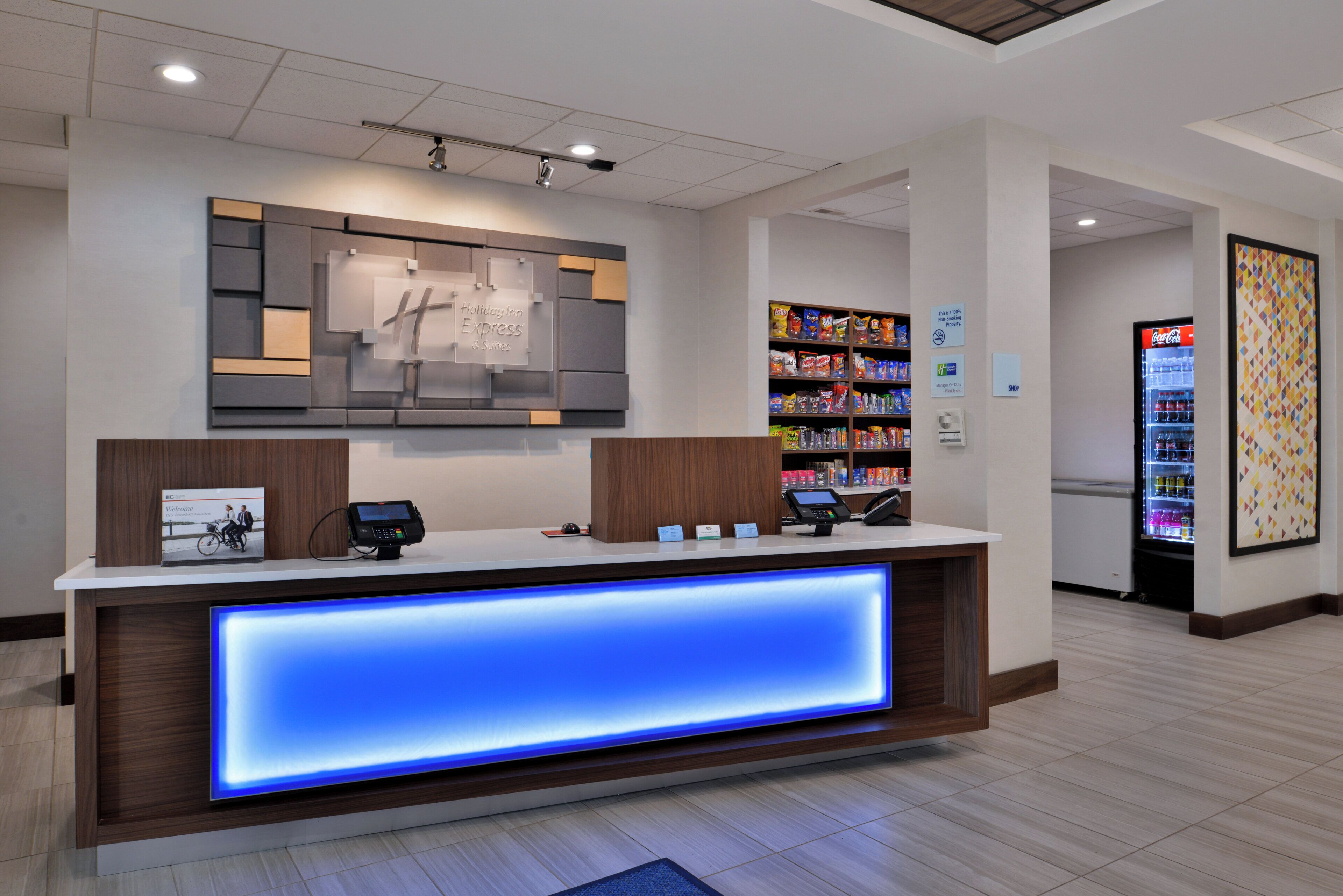 Una recepción de hotel con un panel iluminado en azul, una pantalla digital y una máquina expendedora llena de refrigerios.