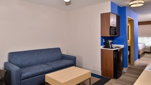 A compact hotel room with a blue sofa, a small wooden coffee table, a kitchenette with a microwave and refrigerator, and a bed in the adjacent room. - Holiday Inn Express & Suites Cincinnati - Mason by IHG (Mason)