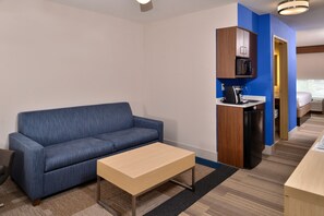 Habitación de hotel compacta con sofá azul, una pequeña mesa de centro de madera, una kitchenette con microondas y refrigerador, y una cama en la habitación contigua.