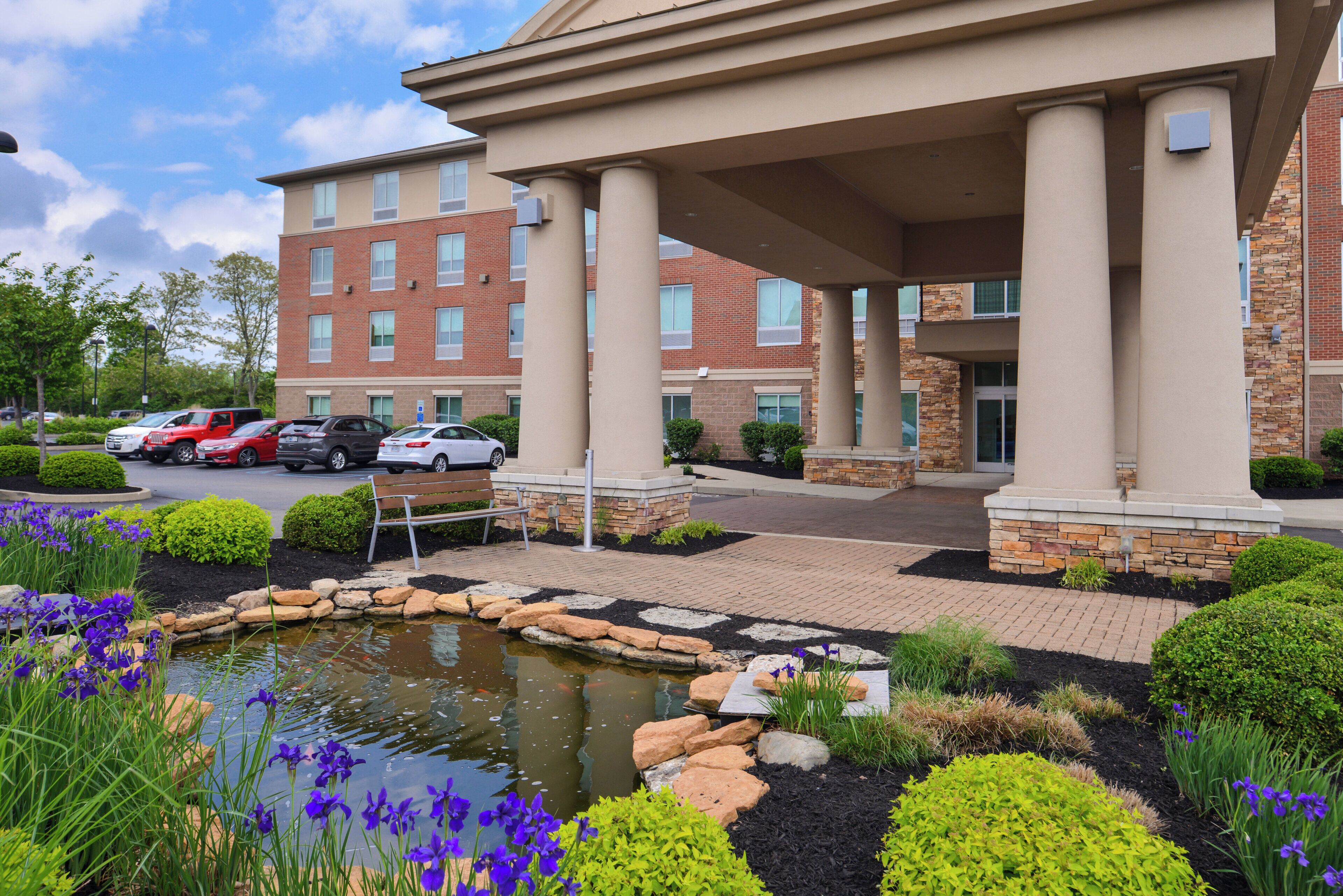 A hotel with a landscaped garden, a pond, and a parking area with cars.