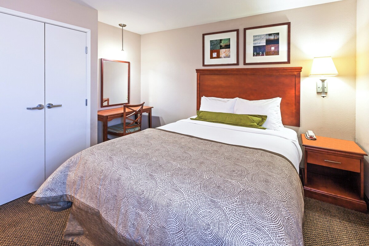 A hotel room with a bed, a desk, a chair, a wardrobe, and two framed pictures on the wall.