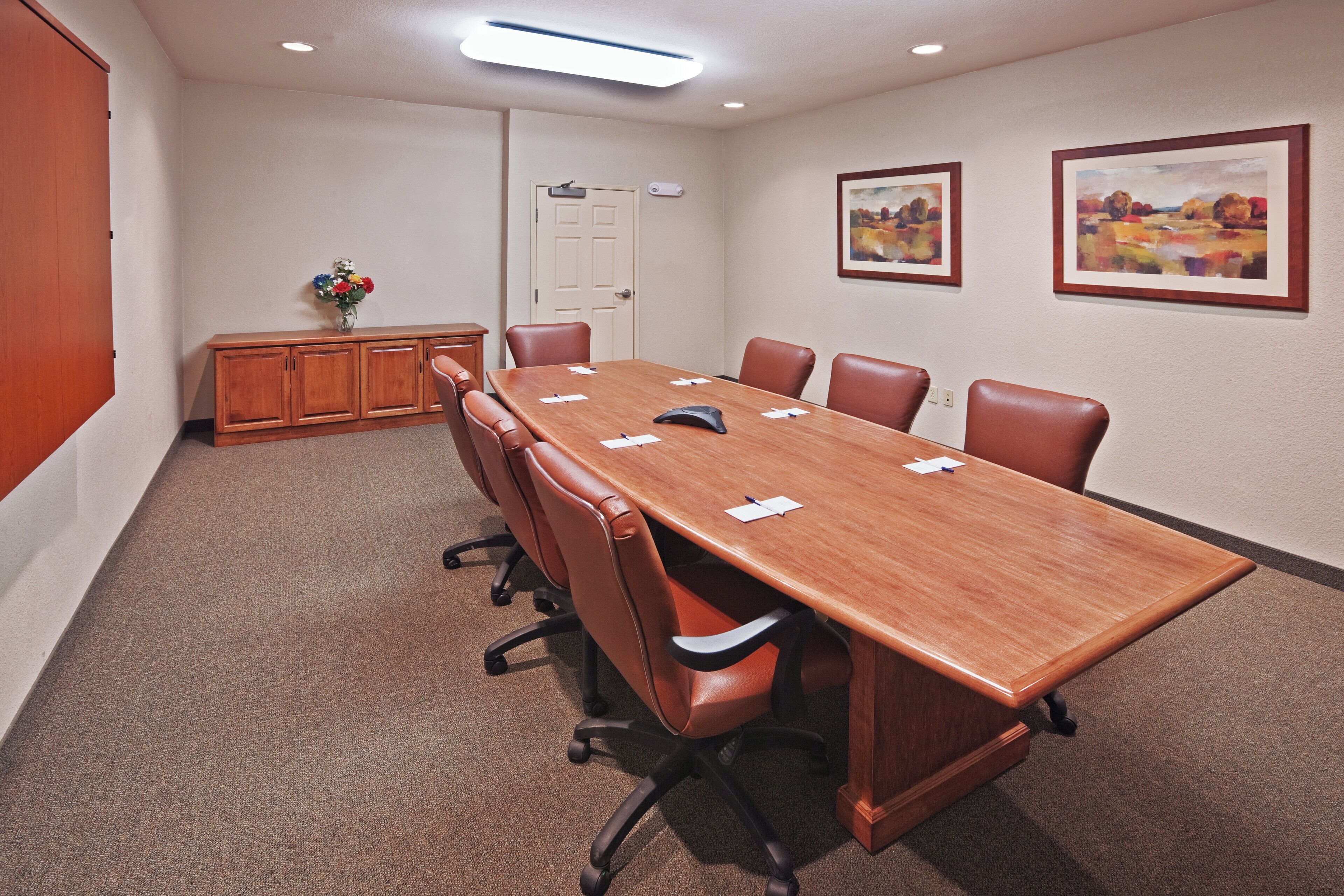 Une salle de conférence avec une longue table, des chaises, un petit meuble et deux tableaux encadrés accrochés au mur.
