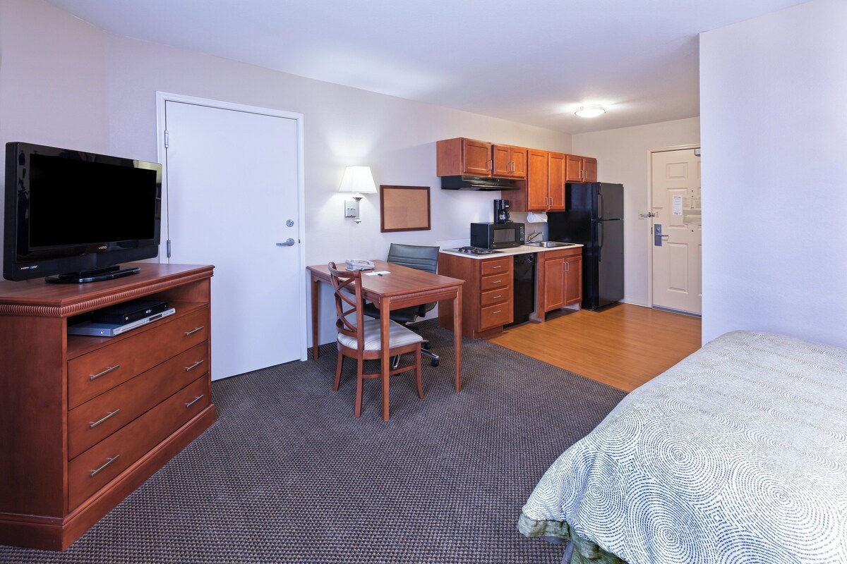 A compact hotel room with a TV, a dining table with chairs, a kitchen area with cabinets and appliances, and a bed with a patterned bedspread.