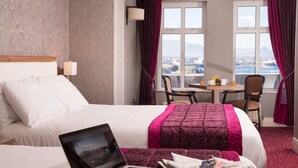 Une chambre d’hôtel avec un lit, un bureau équipé d’un ordinateur portable et une vue sur la ville grâce à de grandes fenêtres.