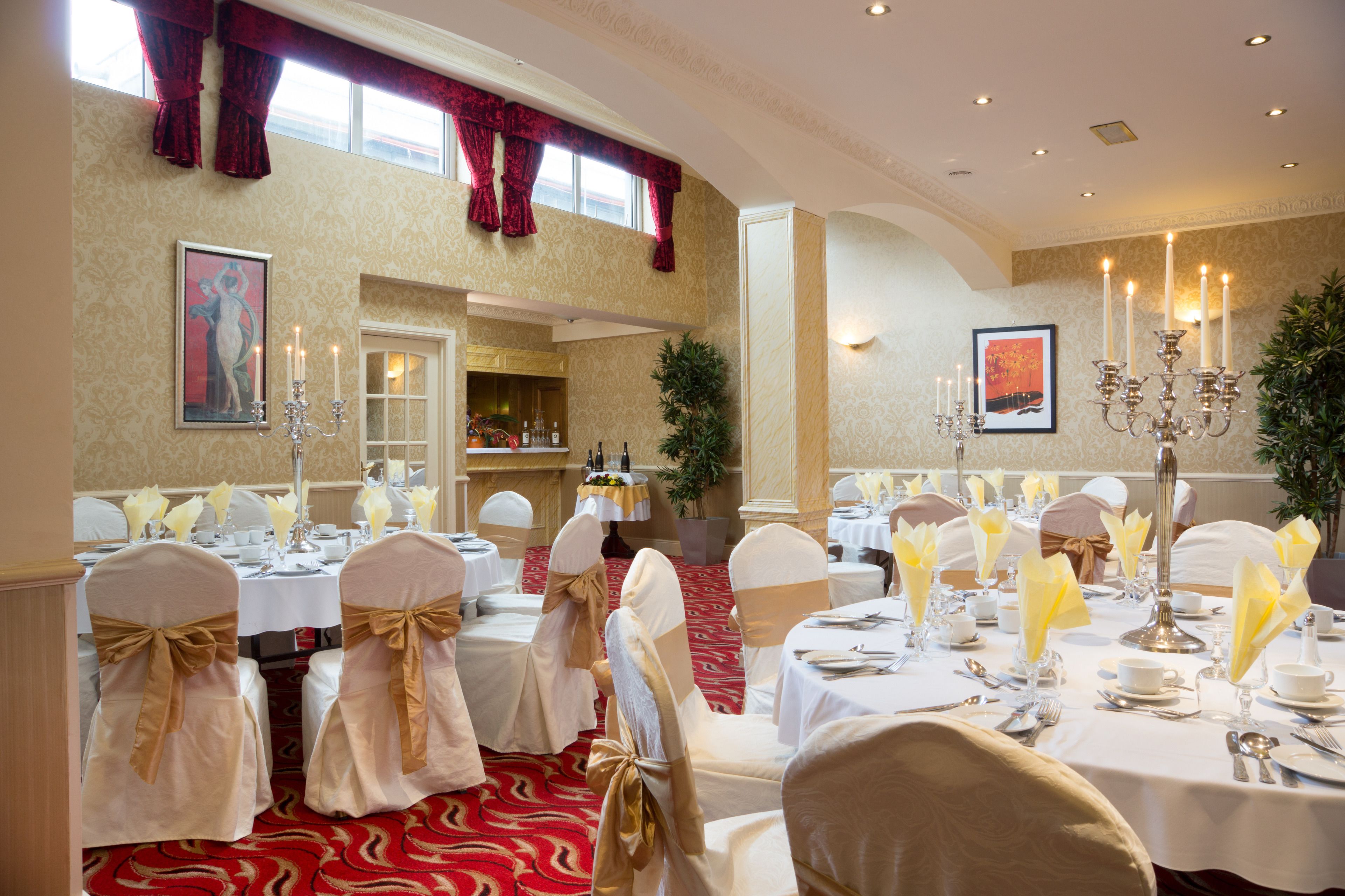 A banquet hall with round tables set for a formal event, featuring white tablecloths, gold sashes on chairs, and candelabras.