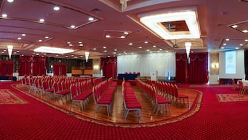 A large conference room with rows of red chairs, a stage with a podium, and a screen displaying a welcome message.