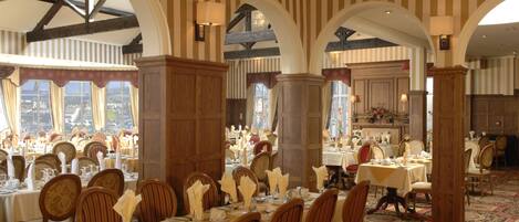 A dining area with multiple tables set for service, wooden chairs, and large windows offering a view.