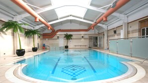 Une piscine intérieure avec un sol carrelé, des palmiers et un plafond haut laissant apparaître les conduits de ventilation.