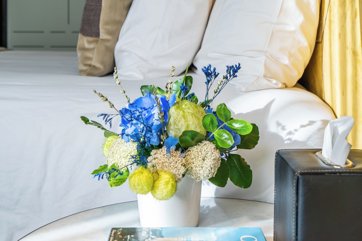 Habitación de hotel ordenada con cama, almohadas, mesita de noche con un libro, un jarrón con flores y una caja de pañuelos.