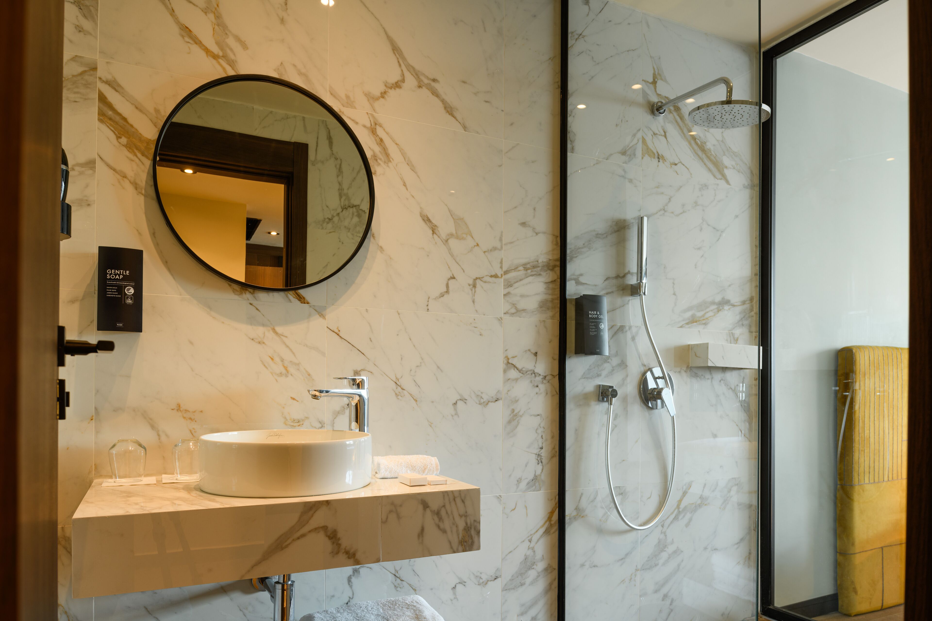 A bathroom with a marble sink, a round mirror, a handheld showerhead, and a glass-enclosed shower.